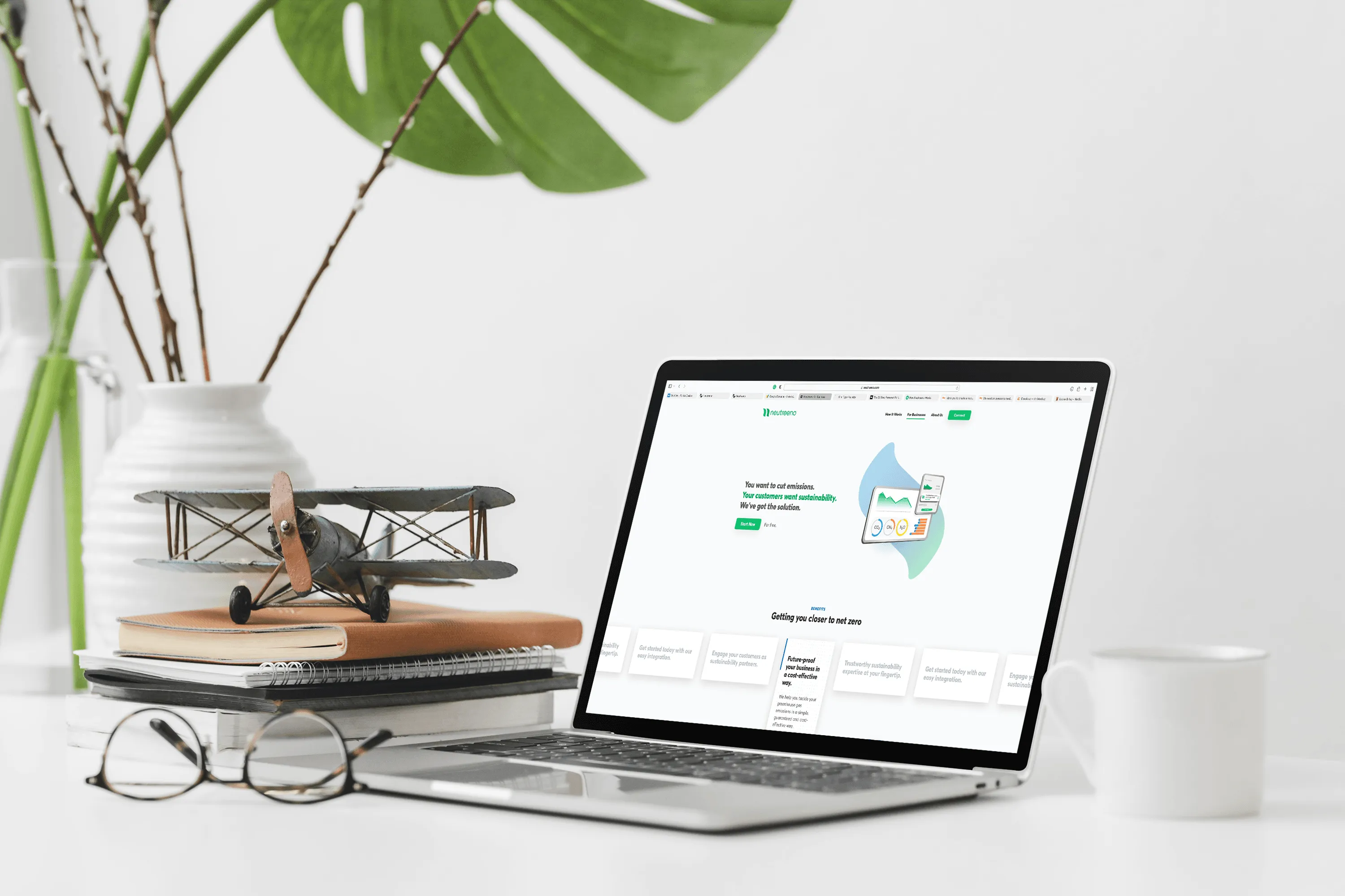 Silver laptop on white desk with wooden airplane model, notebooks, glasses, plant, and white mug.